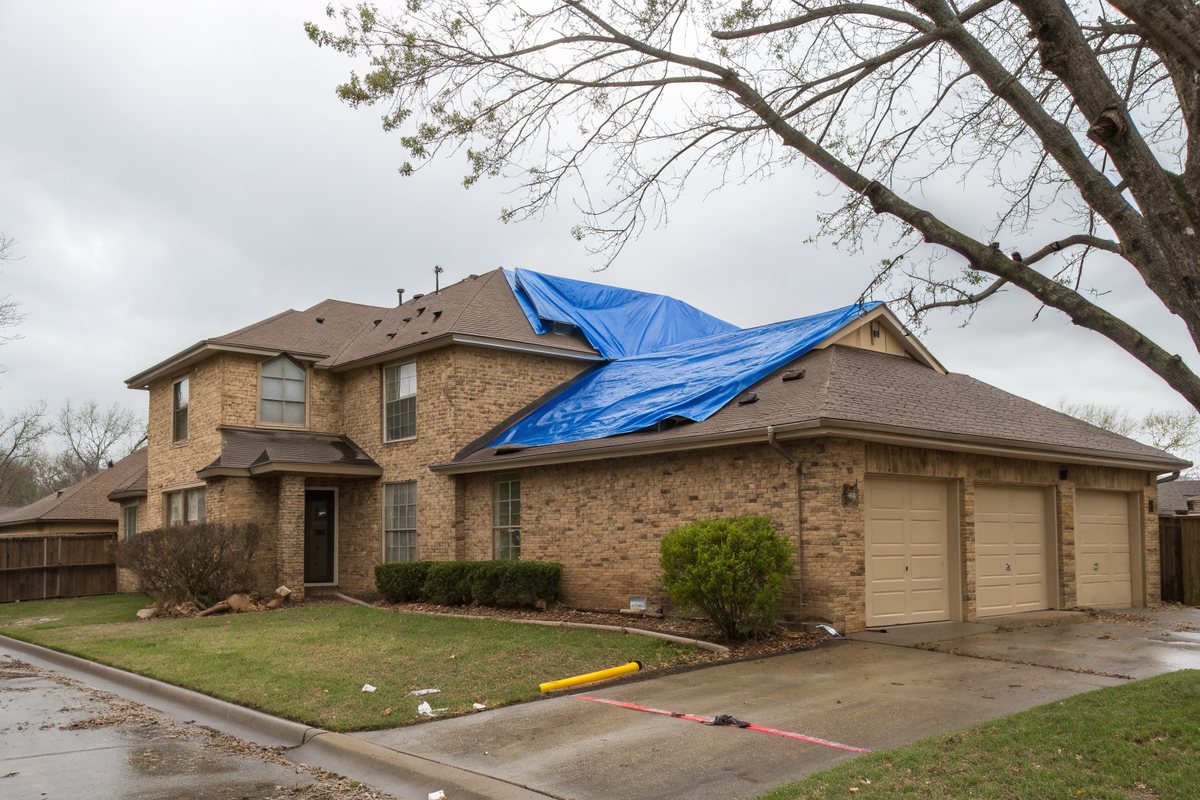 Tan brick two-story home with a three-car garage and a blue tarp spread over part of its brown shingle roof on a gray morning after a storm