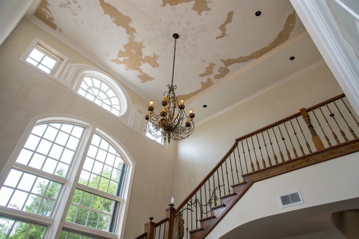 Brown water stains spreading across the white ceiling of a two-story foyer above a brass chandelier, arched windows and a curved stair rail