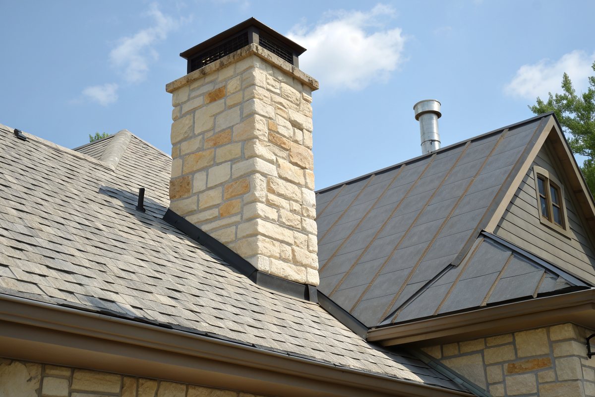 Cream limestone chimney with a dark metal cap and new black metal flashing where it meets a gray shingle roof under a blue sky