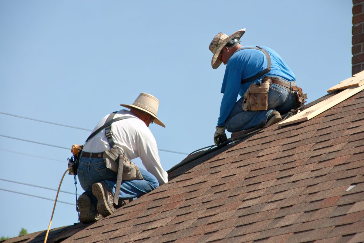 Two roofers in straw sun hats and plain blue and white long-sleeve shirts, seen from behind, kneeling on a brown shingle roof under a clear blue sky